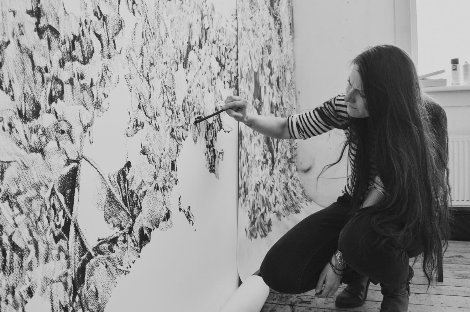 Zwart-witfoto van een vrouw met lang haar die gedetailleerd op een grote rol papier aan de muur tekent met houtskool. Kunstwerk toont complexe patronen en texturen, terwijl persoon knielt op houten vloer in lichte kamer.
