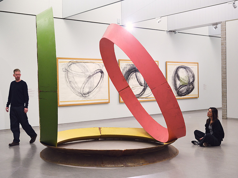 Twee bezoekers bij een kunstwerk, één bezoeker zit op de grond, één bezoeker loopt langs het kunstwerk. 