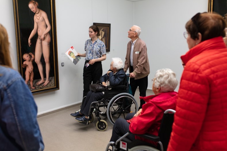 een rondleider toont een schilderij aan een groep ouderen, waarvan twee in rolstoel  | foto: stichting De Zonnebloem, fotograaf Joris Robben