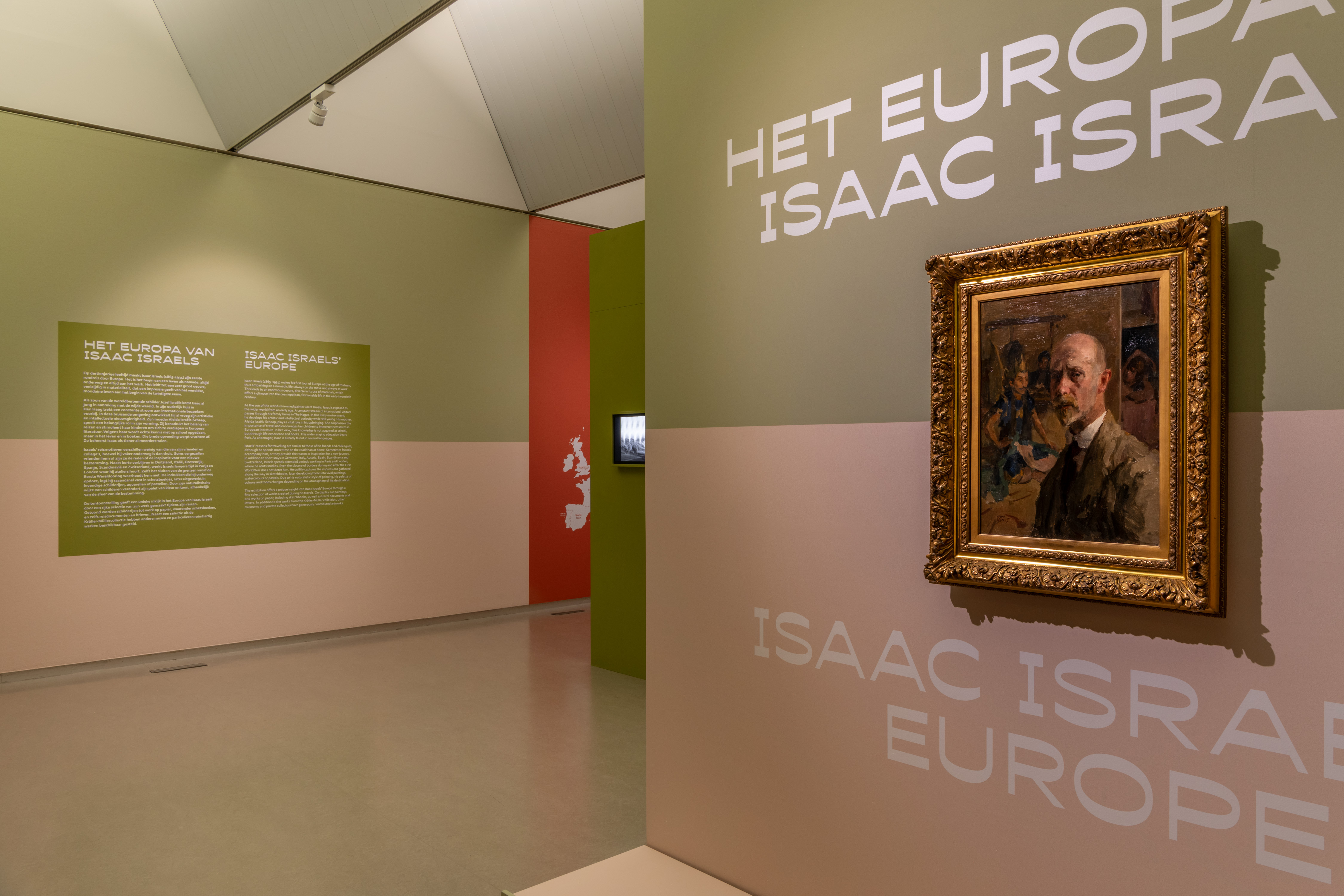 Photo of an exhibition gallery featuring the text &ldquo;The Europe of Isaac Israels&rdquo; on an olive-green wall, with a self-portrait in a gold frame of the (somewhat older) artist in a suit, painted in an impressionist style. In the background, colorful walls in green, pink, and red.