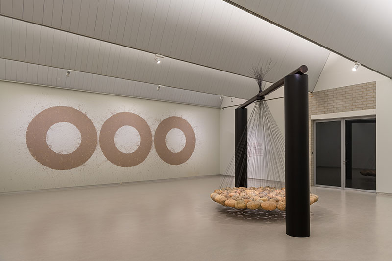 Museumzaal met een installatie van hangende zware keien en een wand met cirkels gemaakt met uitgewreven modder 