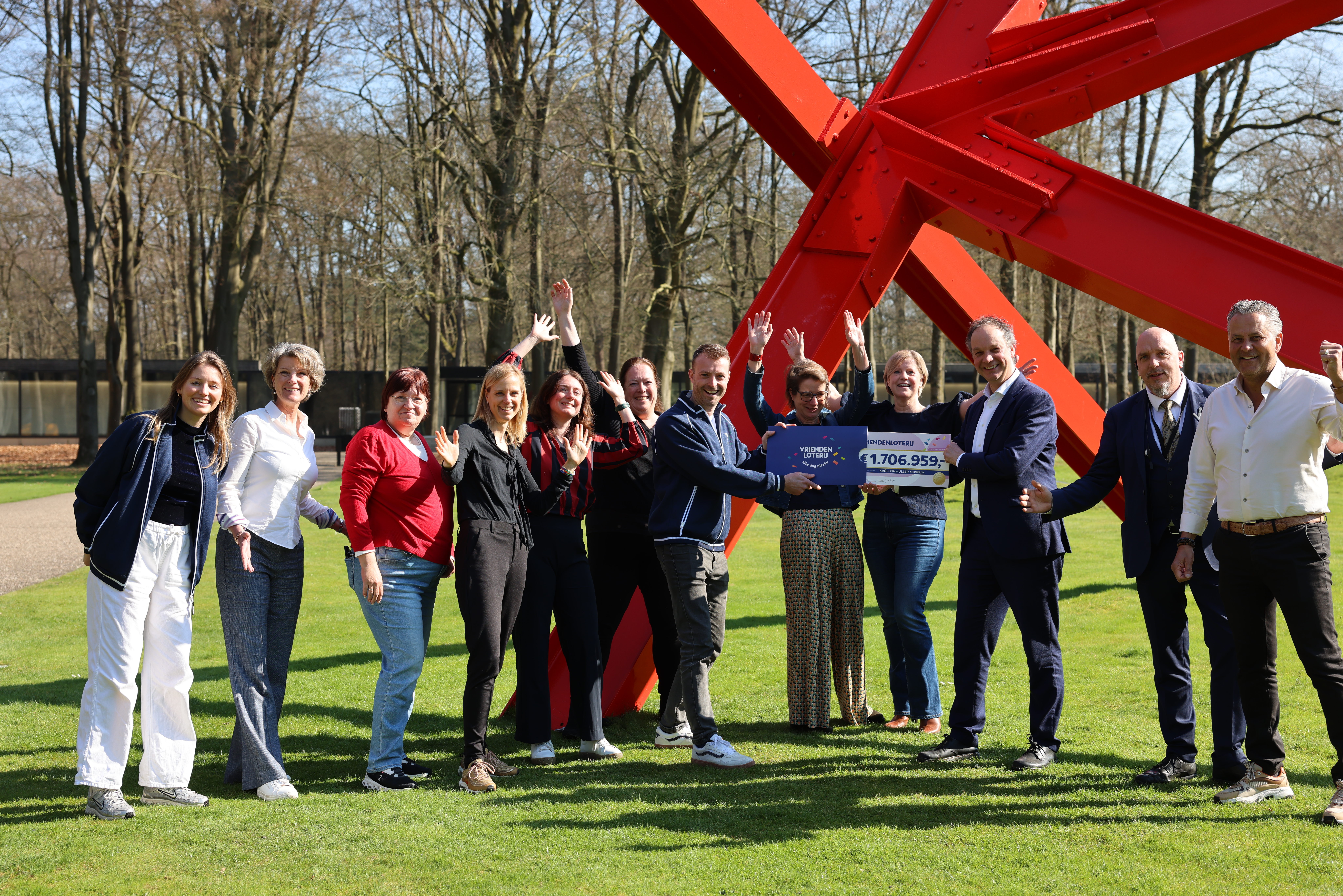 Medewerkers van het museum juichen bij de cheque van de VriendenLoterij. Foto is gemaakt buiten, voor het museum, bij een kunstwerk uit felrode stalen balken